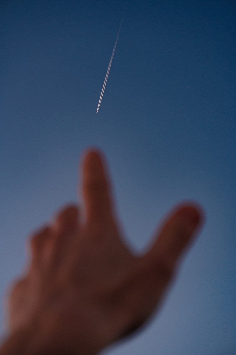 Hand reaching towards a distant airplane contrail